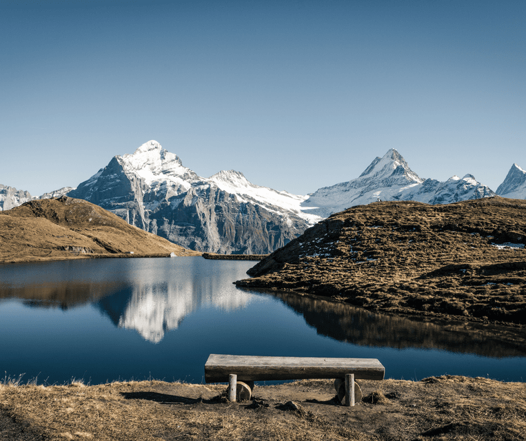 An empty wooden bench on a grassy shore overlooks a calm lake reflecting snow-capped mountains under a clear sky.