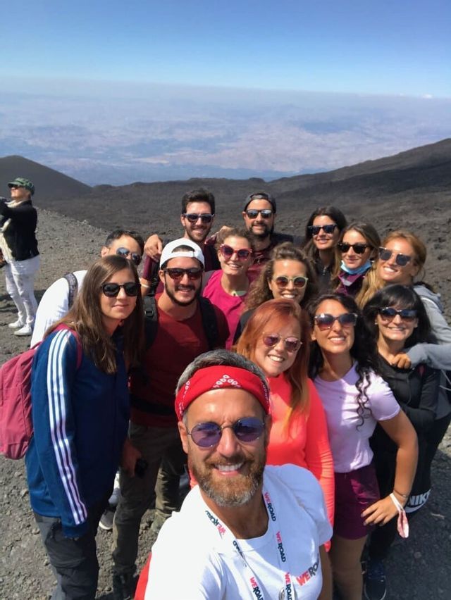 A WeRoad group trip smiles and poses for a selfie on a rocky mountain under a clear blue sky.