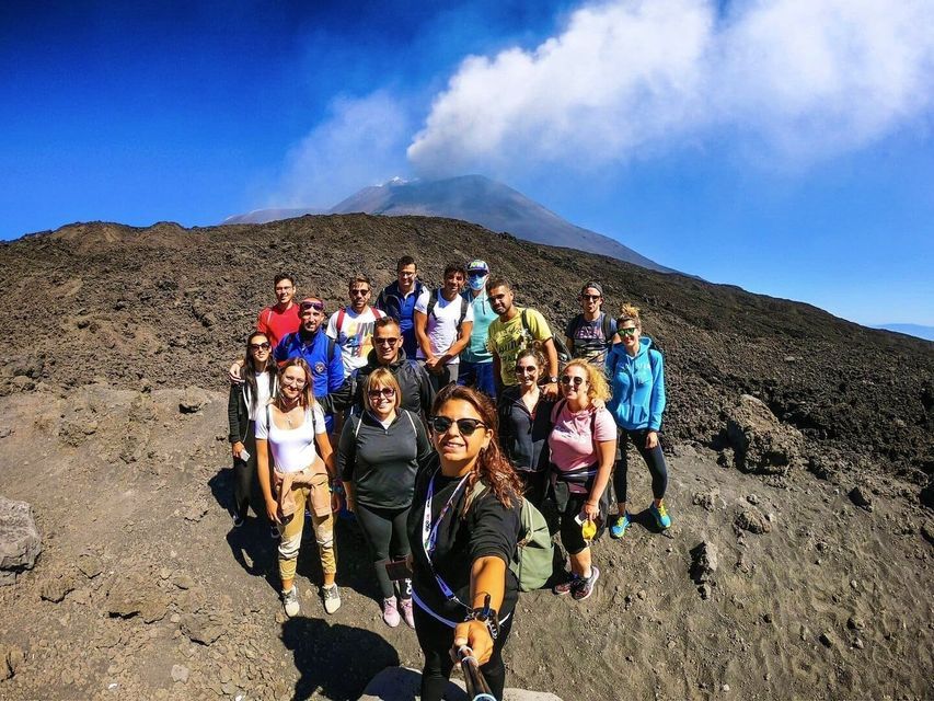 Eine WeRoad-Gruppenreise macht ein Selfie an einem felsigen Berghang, mit einem rauchenden Vulkan im Hintergrund unter blauem Himmel.