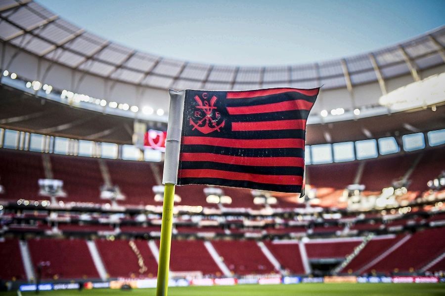 Primo piano di una bandierina d'angolo a strisce rosse e nere con lo stemma del Flamengo, su un campo da calcio, con i seggiolini dello stadio sfocati sullo sfondo.