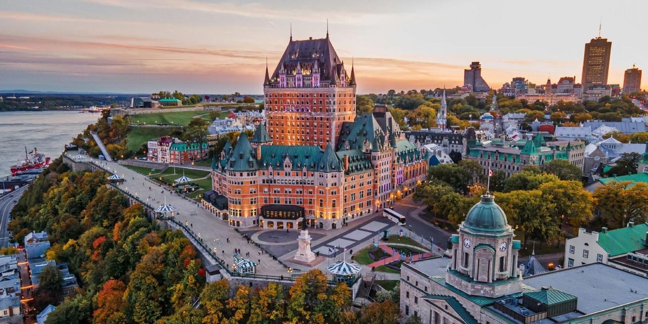 Vista aerea di un magnifico hotel-castello che si affaccia su una città e un fiume al tramonto, circondato da alberi autunnali.