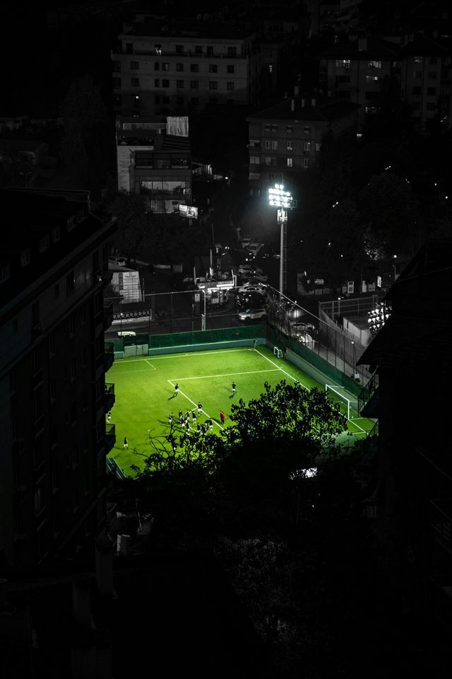 Veduta aerea di una partita di calcio su un campo verde brillantemente illuminato di notte, circondato da edifici scuri della città.