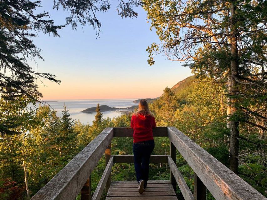 Una persona con una felpa rossa, da un belvedere in legno, ammira una costa boscosa e una baia al tramonto.