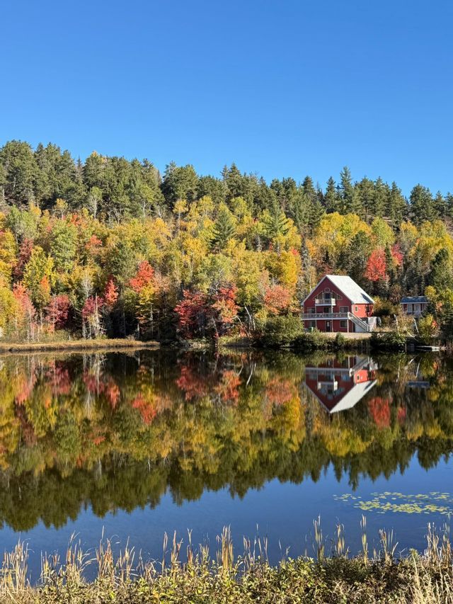 Una casa rossa e una fitta foresta con colori autunnali si riflettono perfettamente nell'acqua calma di un lago.