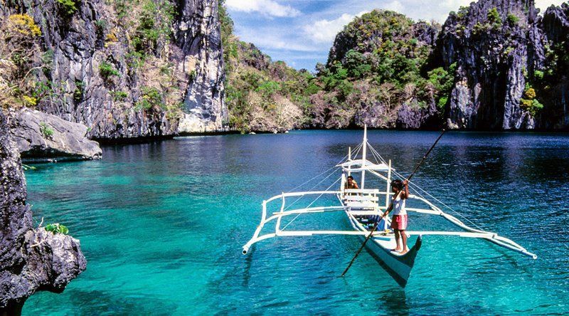 A traditional outrigger boat with two people floats on turquoise water in a calm lagoon between steep, rocky cliffs.