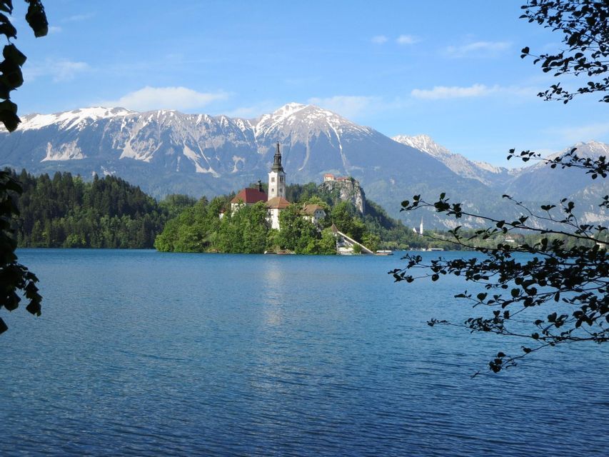 Una chiesa su un'isola boscosa in un lago, con uno sfondo di montagne innevate sotto un cielo blu.