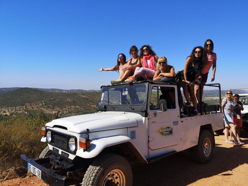 Un viaggio di gruppo WeRoad di donne in posa su una jeep safari bianca su una strada sterrata, con un paesaggio collinare sotto un cielo azzurro e limpido.