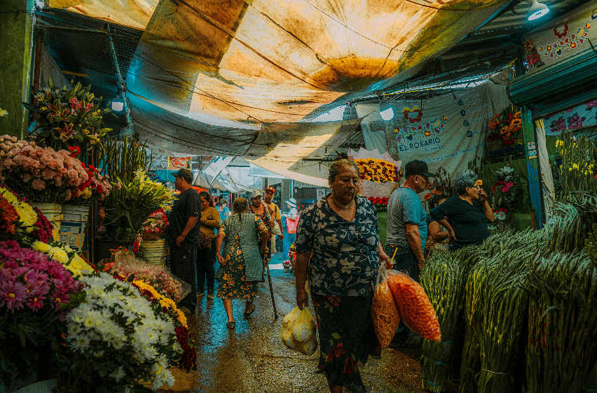 Menschen gehen auf einem nassen Weg durch einen belebten, überdachten Blumenmarkt, umgeben von Ständen voller farbenfroher Blumensträuße.