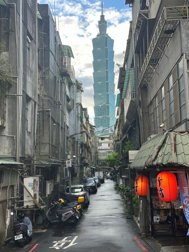 Ein hoher, moderner Glas-Wolkenkratzer, gesehen aus einer engen, nassen Stadtgasse, gesäumt von älteren Gebäuden und geparkten Motorrollern.