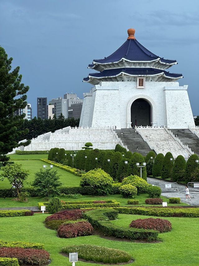 Ein weißes, traditionelles Gebäude mit einem blauen Stufendach liegt hinter einem üppigen, angelegten Garten mit gepflegten Hecken und grünen Rasenflächen.