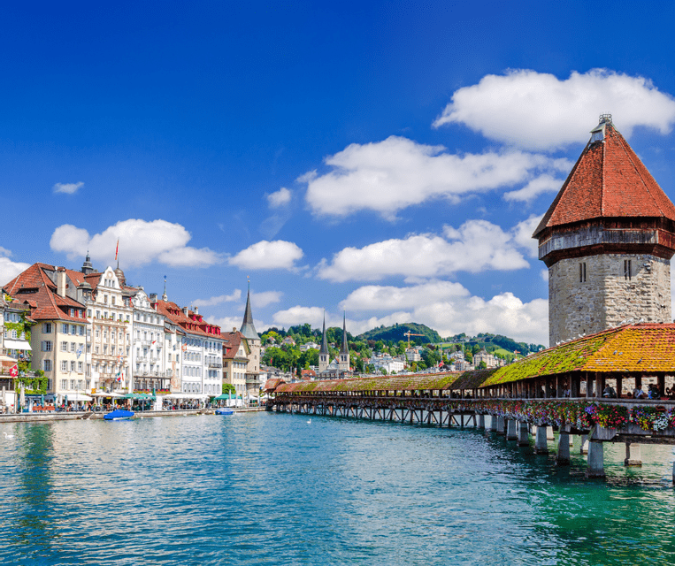 A wooden covered bridge with a stone tower crossing a river next to a historic city waterfront under a blue sky.