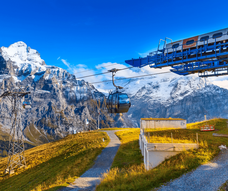 A cable car travels over a green mountain slope, with towering snow-capped peaks visible against a clear blue sky.