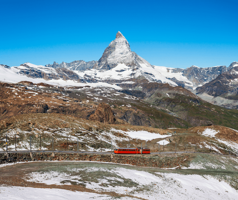 A red train travels on a track across a rocky, partially snow-covered mountainside, with a prominent snow-capped peak in the background.