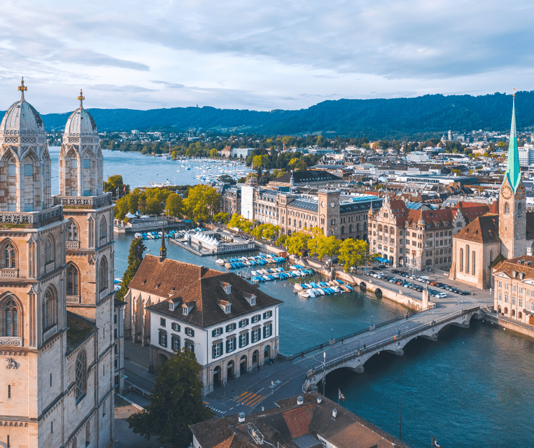 An aerial view of a historic city with two cathedral towers, a river with a bridge, docked boats, and hills in the background.