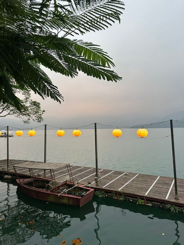 Leuchtende gelbe Papierlaternen hängen über einem Holzsteg, an dem ein kleines Boot auf einem ruhigen See vertäut ist.