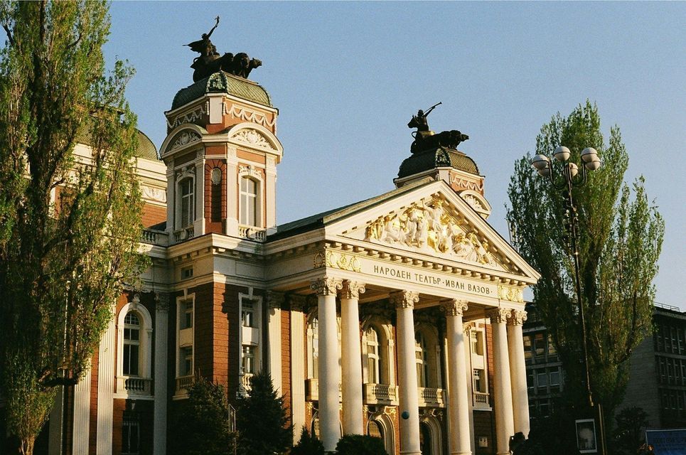 La fachada iluminada por el sol de un edificio ornamental de estilo neoclásico con grandes columnas blancas, un frontón dorado y estatuas en sus torres.