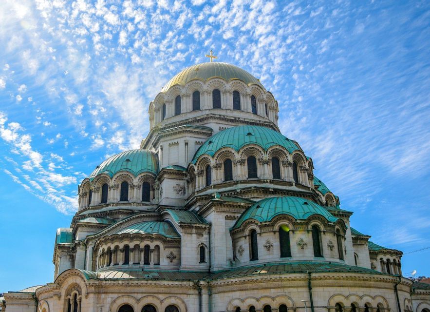 Una gran catedral con una cúpula central dorada y cúpulas turquesas más pequeñas contra un cielo azul brillante con nubes ligeras.