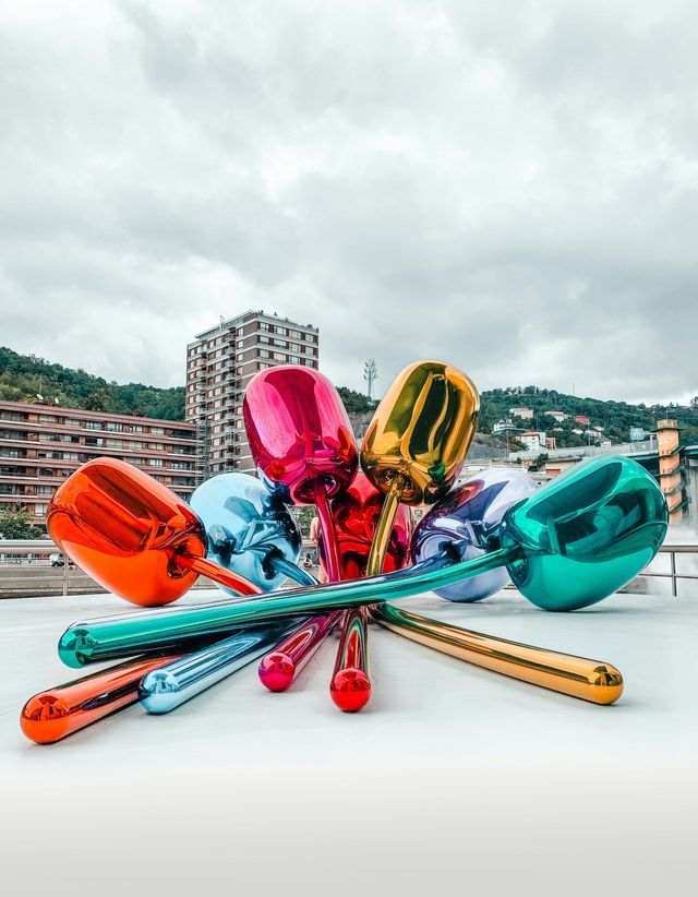 Una grande scultura colorata di tulipani metallici giace su una piattaforma bianca in una città sotto un cielo coperto.
