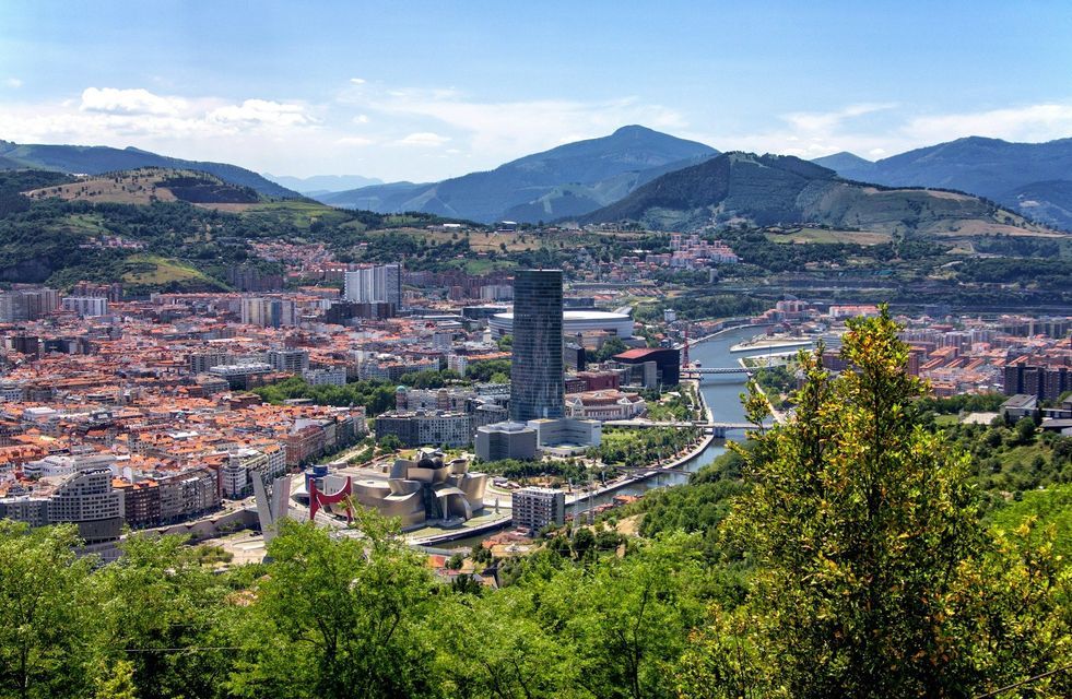 Una veduta panoramica di una grande città con un fiume serpeggiante, incastonata tra dolci colline verdi e montagne sotto un cielo blu.