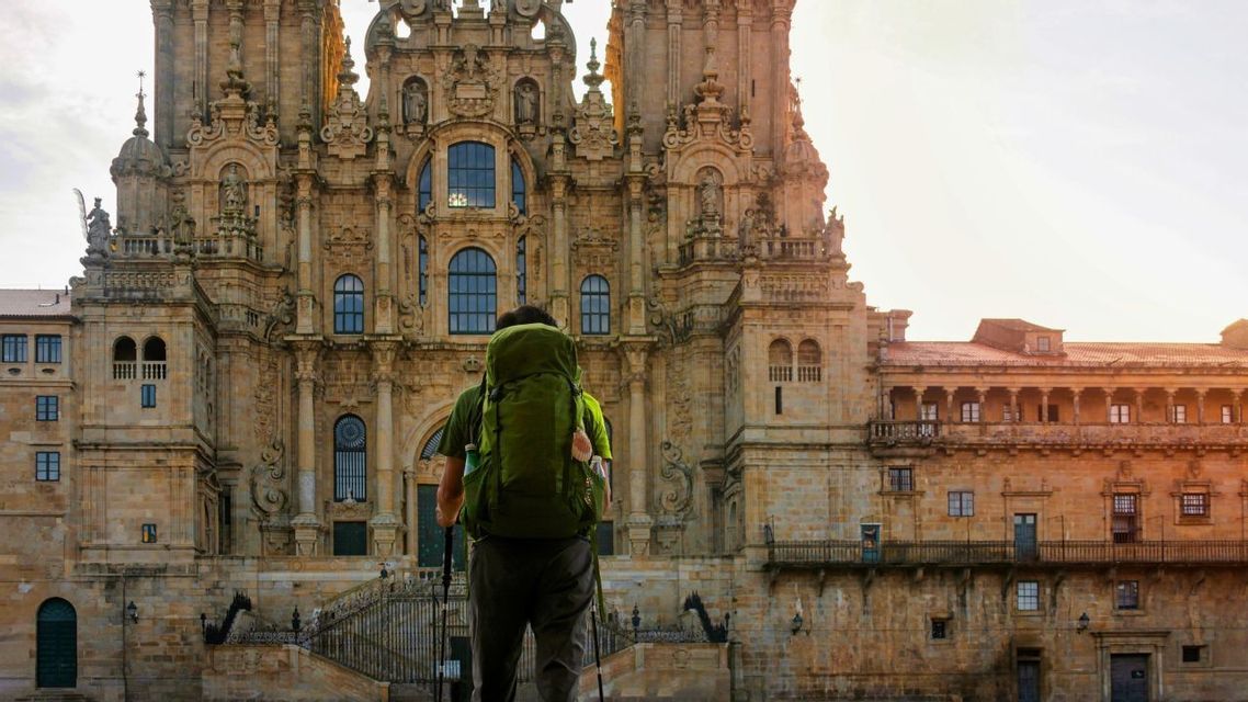 Ein Reisender mit großem grünem Rucksack und Trekkingstöcken blickt zur verzierten Fassade einer historischen Steinkathedrale auf.