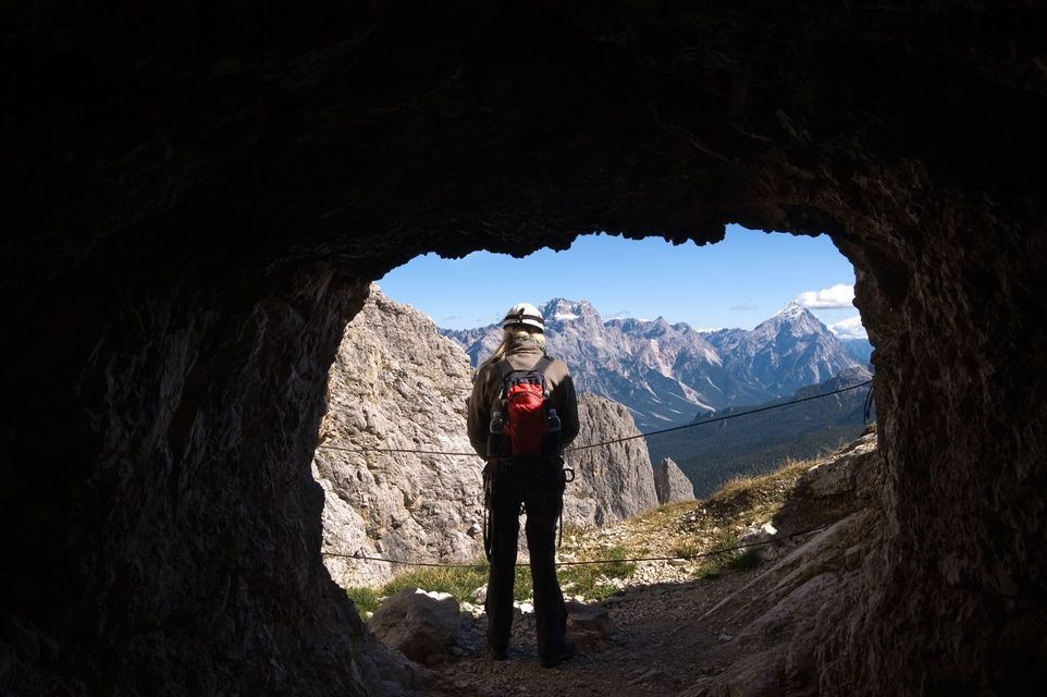 Un excursionista con casco y mochila roja está en la boca de una cueva, contemplando una vasta cordillera iluminada por el sol.