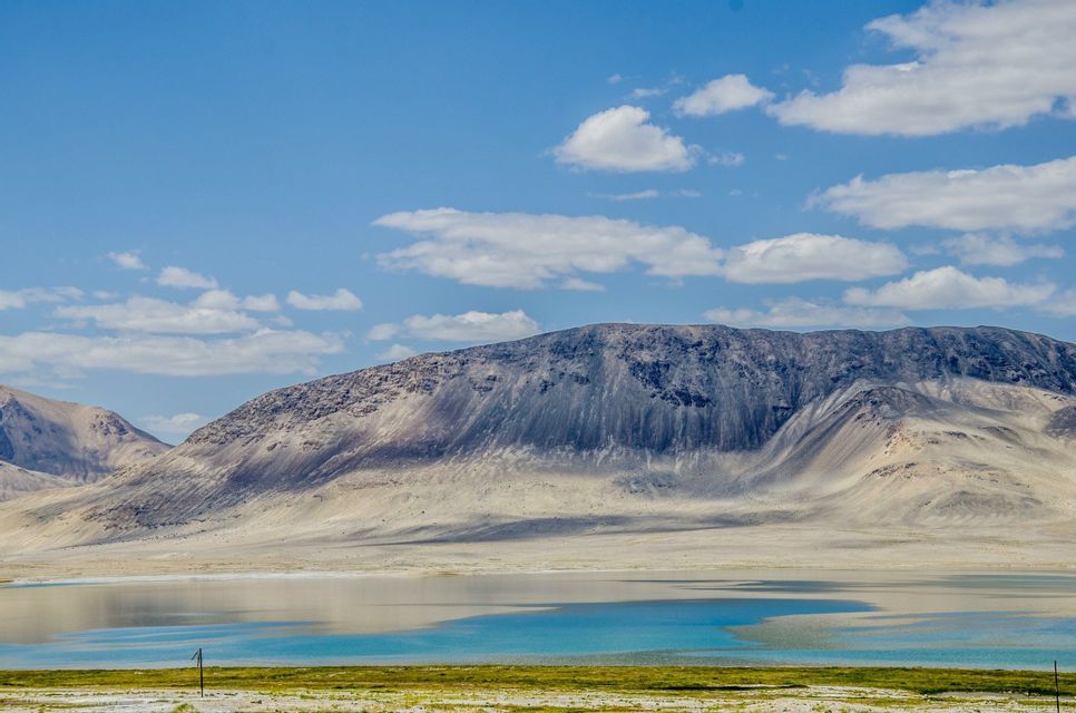 Ein ruhiger türkisfarbener See liegt am Fuße eines großen, kargen Berges unter einem teilweise bewölkten blauen Himmel.