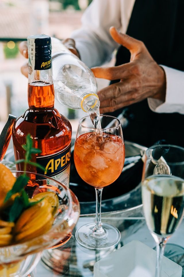 Les mains d'un barman versent un liquide transparent d'une bouteille dans un verre à vin avec des glaçons, à côté d'une bouteille d'Aperol.