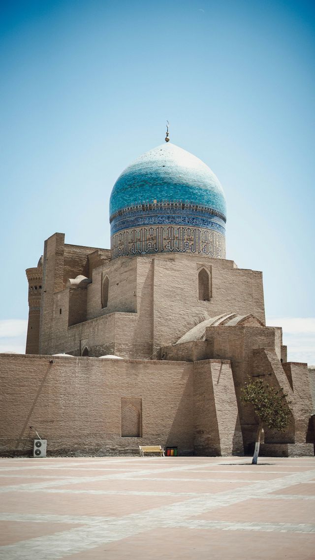 Un grande edificio in mattoni con una cupola decorata di piastrelle blu si erge in una piazza pavimentata sotto un cielo azzurro e limpido.