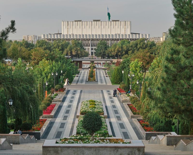 Une large allée symétrique de parc, bordée d'arbres et de parterres de fleurs colorés, mène vers un grand bâtiment moderne et blanc au loin.