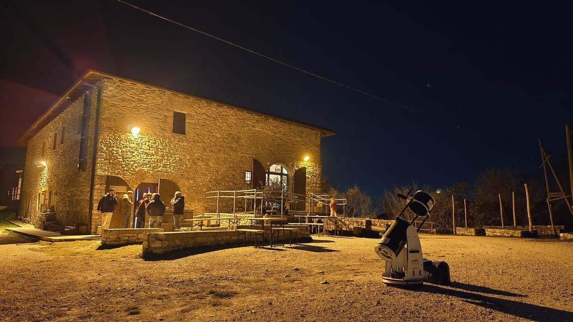 Un grande telescopio si erge in una radura ghiaiosa di notte, puntato verso il cielo, con un gruppo WeRoad riunito fuori da un edificio in pietra illuminato.
