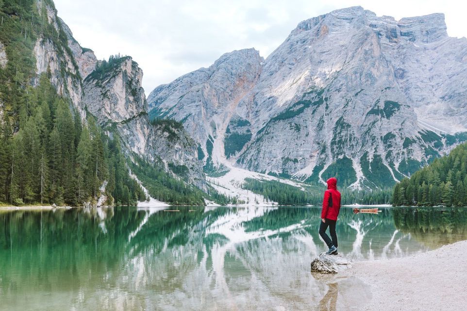 A person in a red jacket stands on a rock by a lake, looking at mountains reflected in the water.