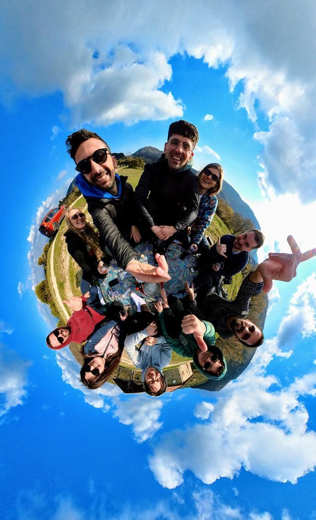 Un viaggio di gruppo WeRoad che sorride per un selfie a 360 gradi effetto 'piccolo pianeta' in un paesaggio verde e collinare sotto un cielo azzurro.