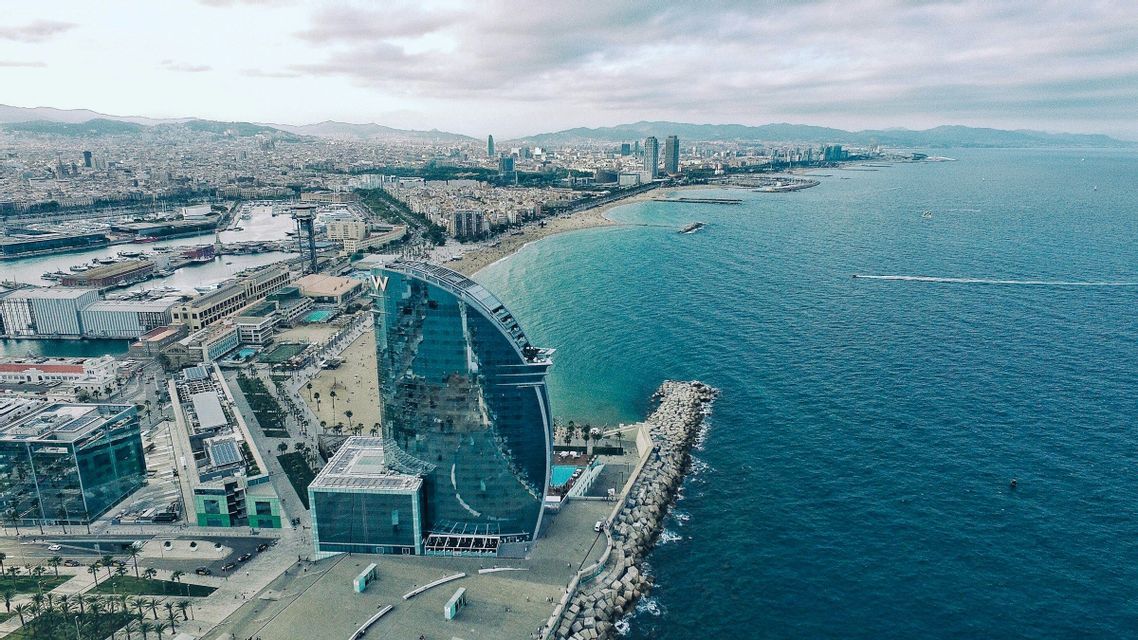 Vista aerea di una città costiera con un hotel di vetro a forma di vela sul lungomare, accanto a una lunga spiaggia sabbiosa.