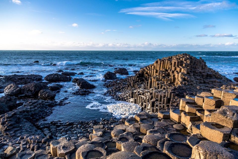 Colonne esagonali di basalto incastrate formano un paesaggio costiero, con le onde dell'oceano che lambiscono la riva sotto un cielo azzurro.