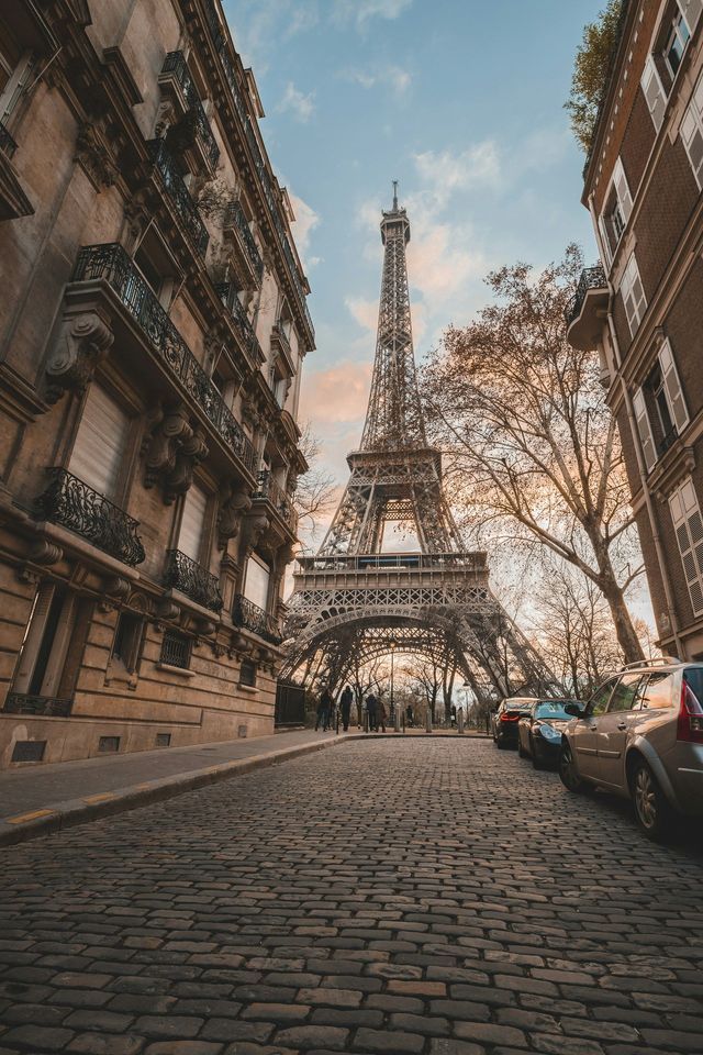 Una vista dal basso della Torre Eiffel da una strada acciottolata, incorniciata da edifici tradizionali e alberi sotto un cielo nuvoloso.