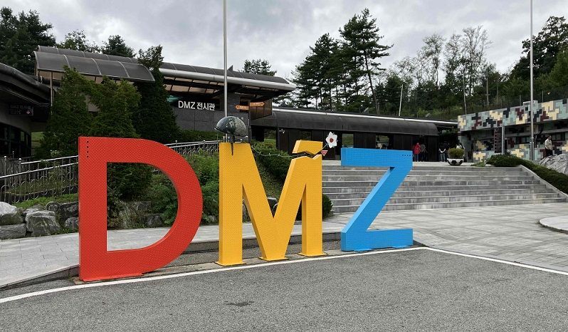 Grandi lettere colorate che compongono la scritta DMZ si ergono di fronte a un padiglione espositivo, con un fiore bianco che spunta dalla 'M' spezzata.