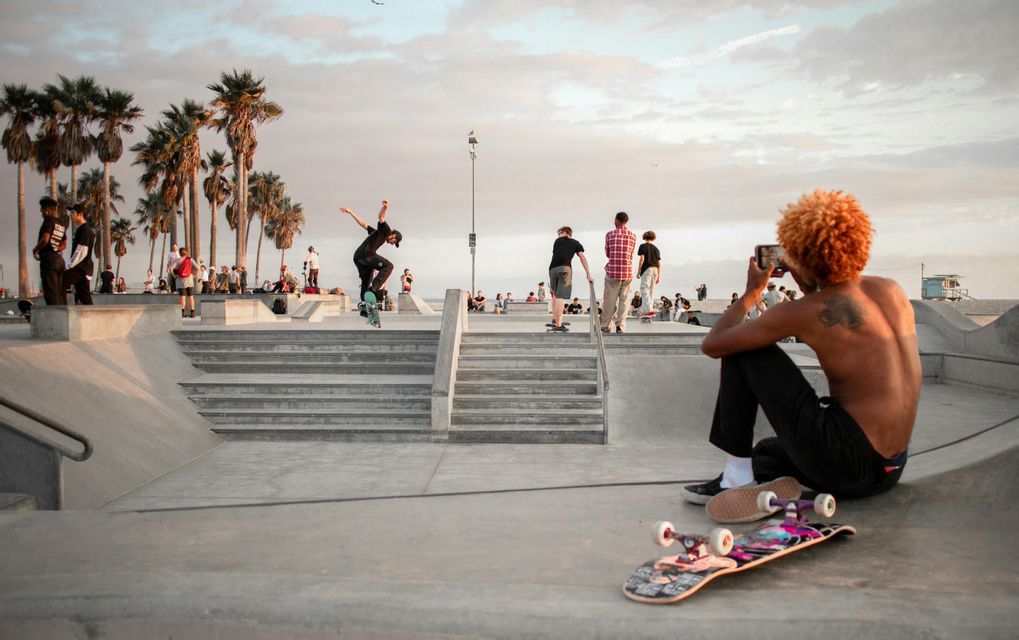 Una persona riprende con il telefono mentre un altro skater esegue un trick a mezz'aria in uno skatepark di cemento con palme.