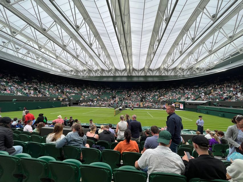 Una grande folla siede sui seggiolini verdi dello stadio guardando una partita di tennis su un campo in erba sotto un grande tetto retrattile.