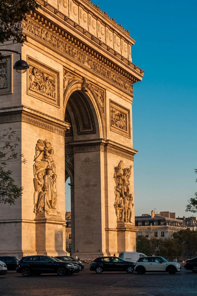 Il fianco di un maestoso arco monumentale è illuminato dalla luce dorata del sole, con auto sulla strada sottostante e un cielo azzurro limpido.
