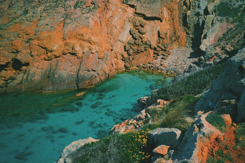 Vista dall'alto di una caletta rocciosa con acqua cristallina turchese, circondata da ripide scogliere rosso-arancio e vegetazione verde.