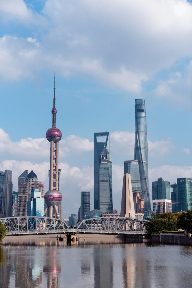 Lo skyline moderno di Shanghai, con la Torre della Perla Orientale, si erge sopra un fiume e un ponte d'acciaio sotto un cielo blu con nuvole bianche.