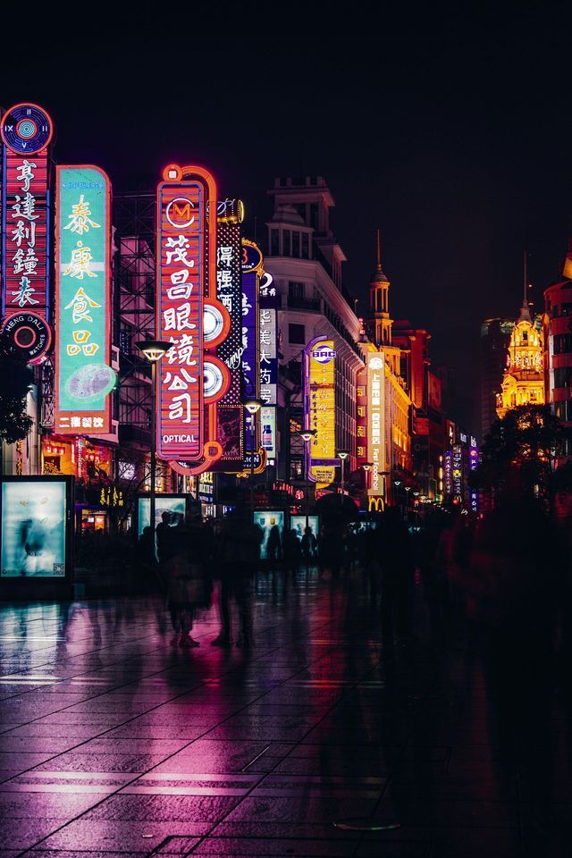 Di notte, una strada cittadina bagnata riflette il bagliore di vivaci insegne al neon verticali con caratteri asiatici, mentre la gente passeggia.