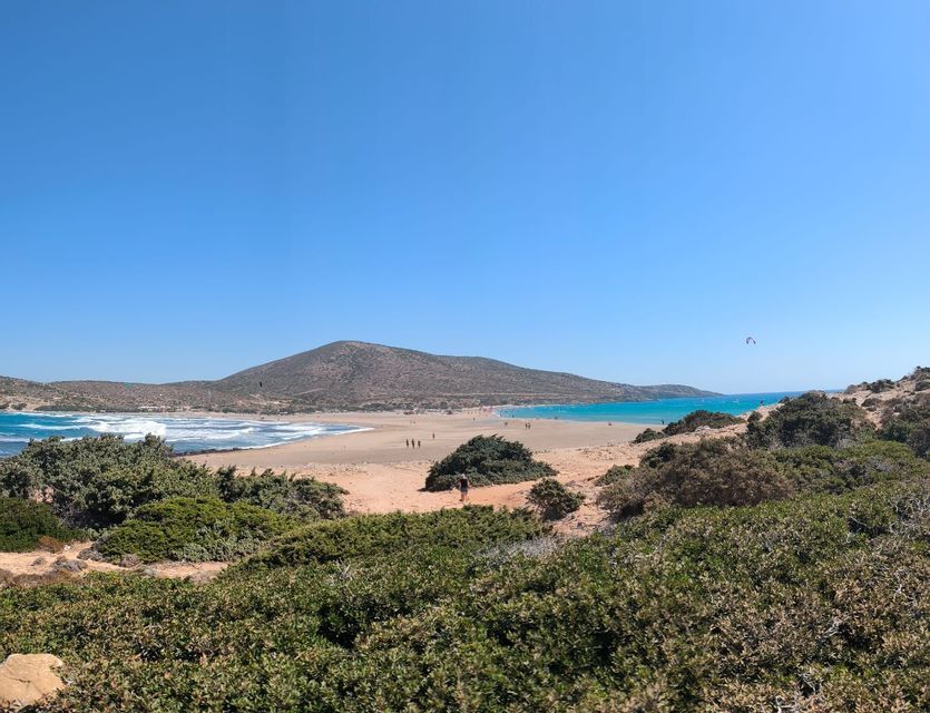 Una vista panoramica di un'ampia spiaggia sabbiosa con acqua turchese e onde, vista da una collina coperta da arbusti verdi sotto un cielo azzurro chiaro.