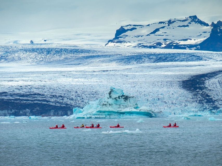 Un viaggio di gruppo WeRoad pagaia in kayak rossi attraverso acque ghiacciate di fronte a un enorme ghiacciaio e montagne innevate.
