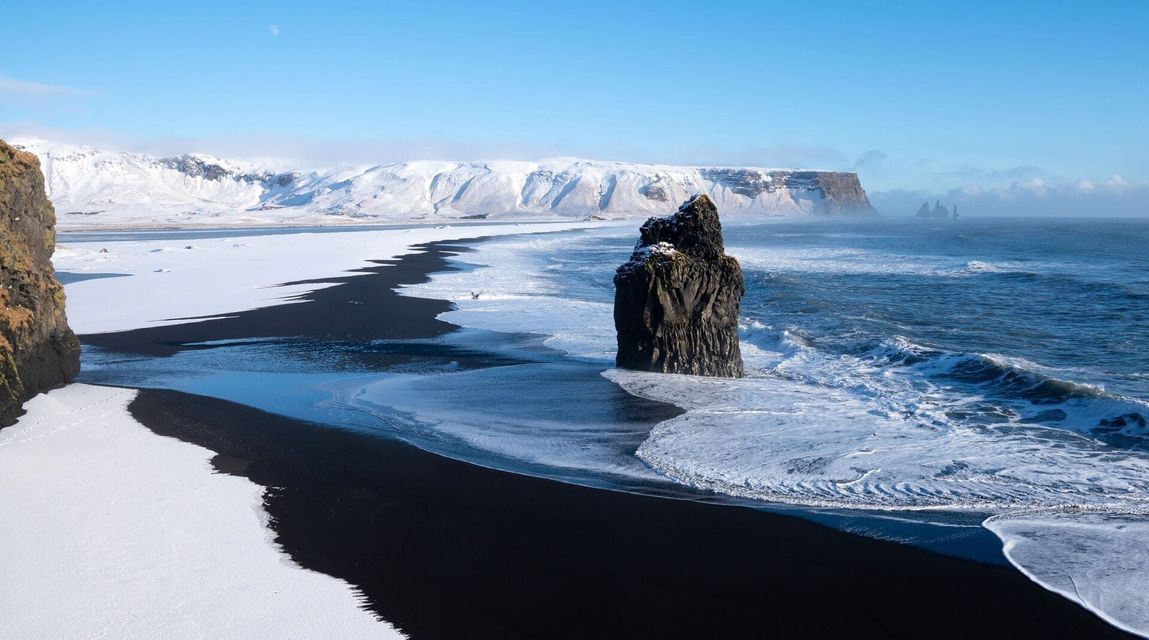 Un imponente farallón marino se erige en el agua frente a una playa de arena negra, parcialmente cubierta de nieve, con montañas nevadas al fondo.