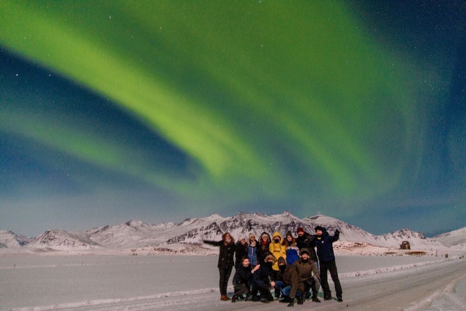 Un grupo de WeRoad posa para una foto bajo la aurora boreal verde en un paisaje nevado con montañas.
