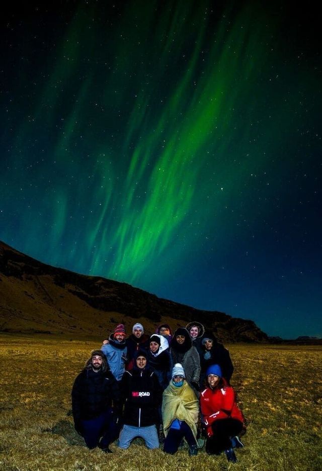Un viaje en grupo de WeRoad posando para una foto en un campo de noche bajo la aurora boreal verde y un cielo estrellado.