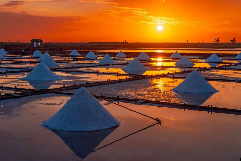 Pyramidenförmige Haufen weißen Salzes liegen in flachen Verdunstungsbecken und spiegeln einen leuchtend orangefarbenen Sonnenuntergang wider.