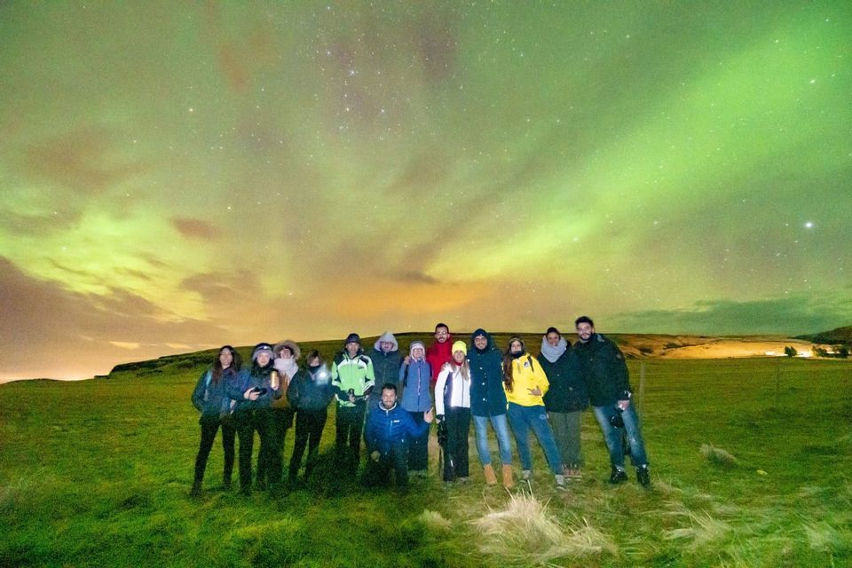 A WeRoad group trip posing for a photo on a grassy hill under the green Northern Lights and a starry night sky.