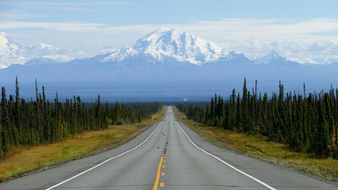 A straight paved road stretches through a forest of evergreens towards a large, snow-capped mountain range in the distance.